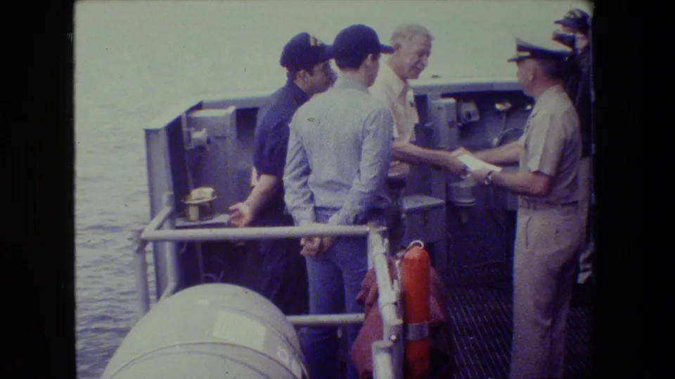 A man in dark clothing and cap stands near another man in a light shirt while a uniformed officer presents or reads from a document on the ship deck.