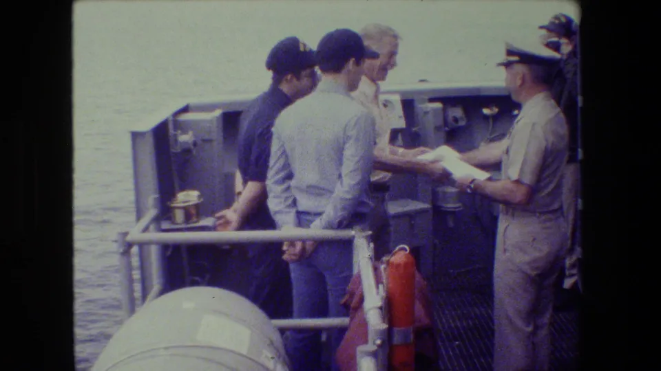 The final frame holds on the shipboard group with the officer and papers at right, men in caps at center, and water beyond the deck railings.