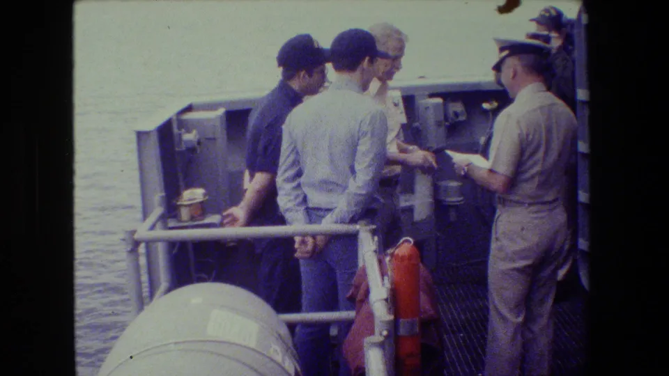 In archival 8mm footage identified as 1981 naval-base material, several adult men gather on a ship deck as a uniformed officer appears to review a paper.