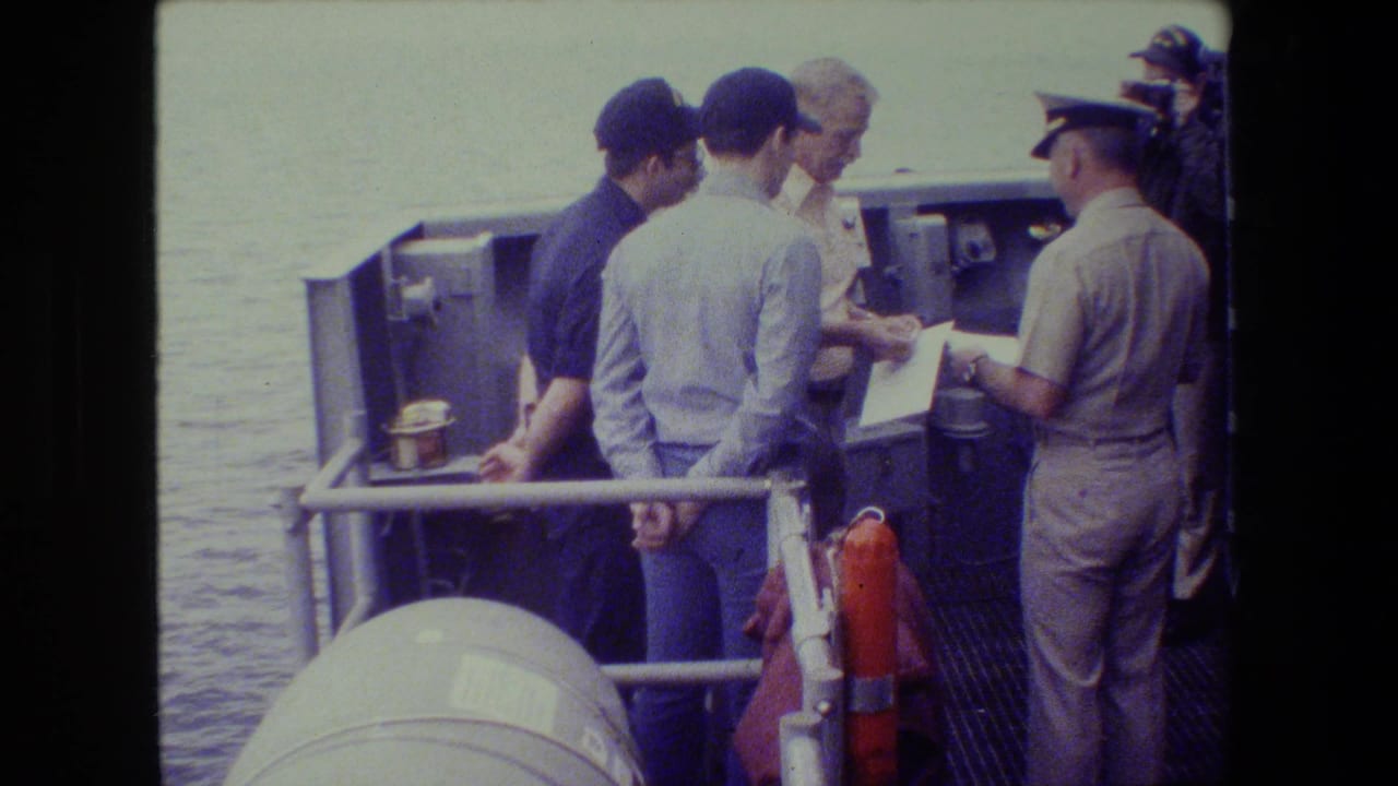 A light-uniformed naval officer stands near deck equipment with a paper in hand while several men watch, with water visible beyond the railing.