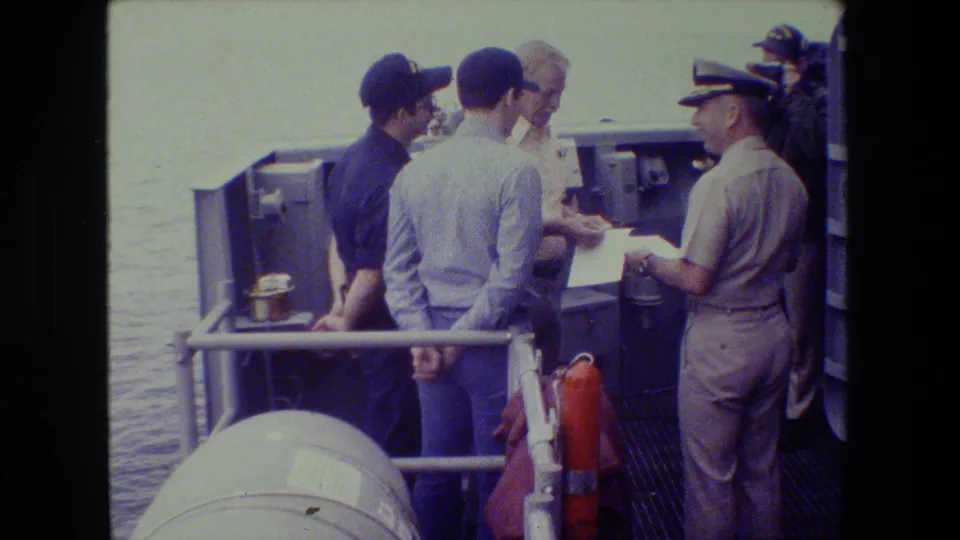 The group remains clustered around paperwork on the ship deck, with a uniformed officer facing the men and water visible behind the rail.