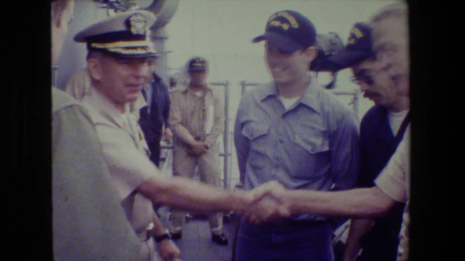 In 1981 archival 8mm footage, a Navy officer in a khaki uniform and peaked cap appears to shake hands with a civilian man on a ship deck as another sailor in blue looks on.