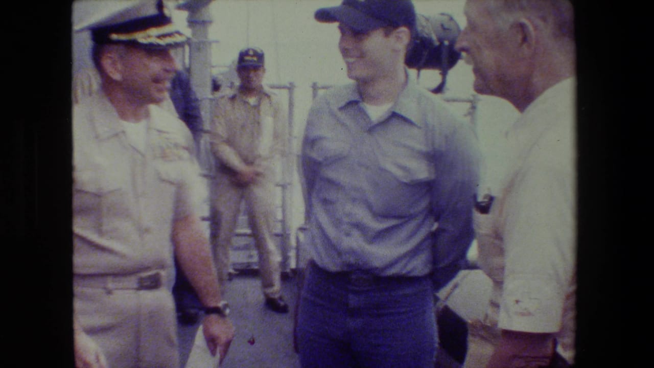 A Navy officer in a light khaki uniform stands smiling beside a sailor or worker in a blue shirt and cap, with an older civilian man at the edge of the shipboard group.