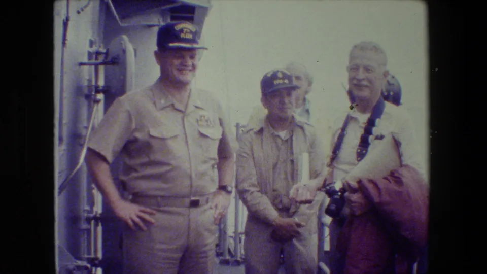A khaki-uniformed Navy officer stands on deck with hands on hips near another uniformed man and an older civilian holding papers and camera equipment.