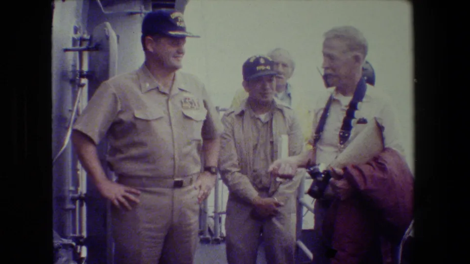 A Navy officer in khaki stands with two men on a ship deck, including a cap-wearing uniformed man and an older civilian with camera gear and papers.