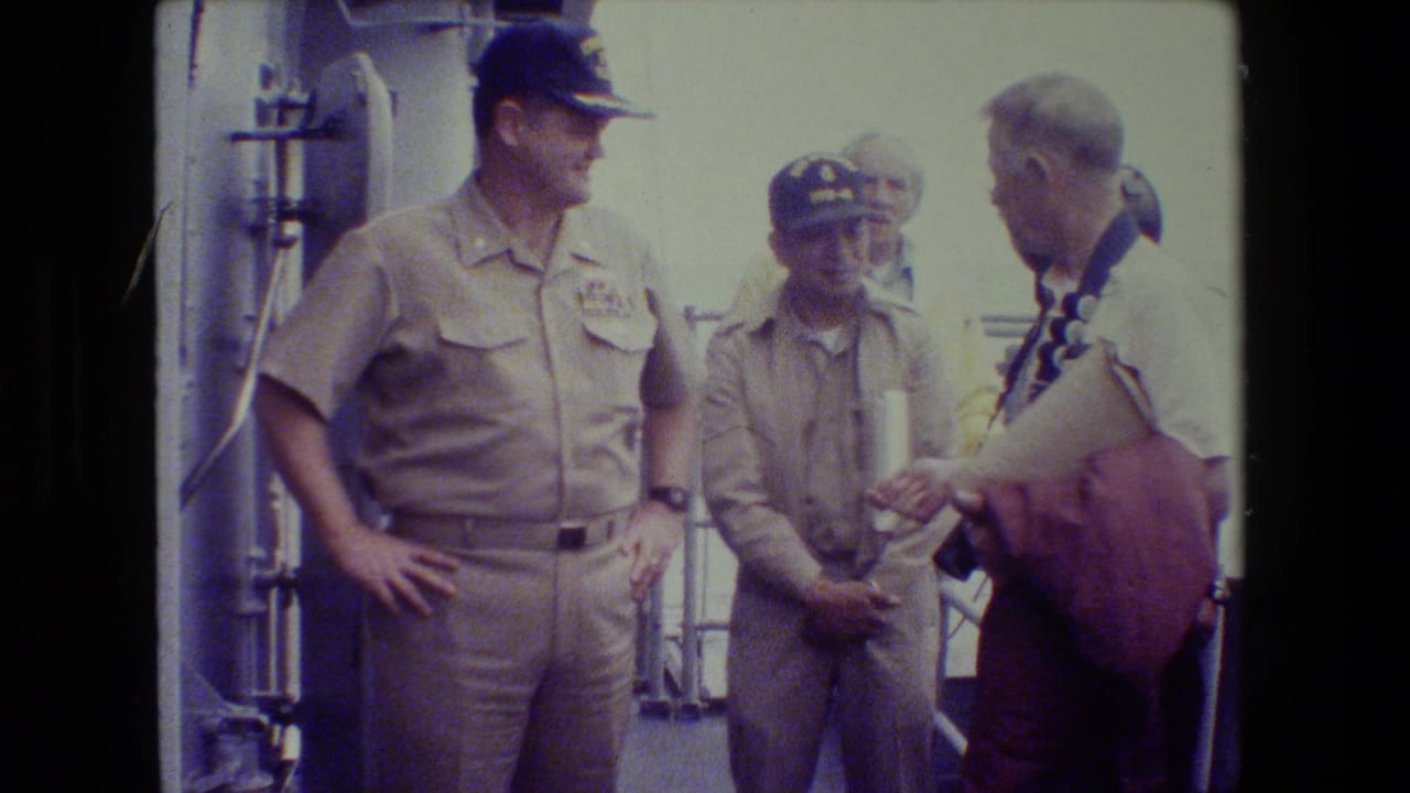 A khaki-uniformed naval officer stands with hands on hips beside another uniformed man and a civilian visitor holding camera gear on a ship deck.