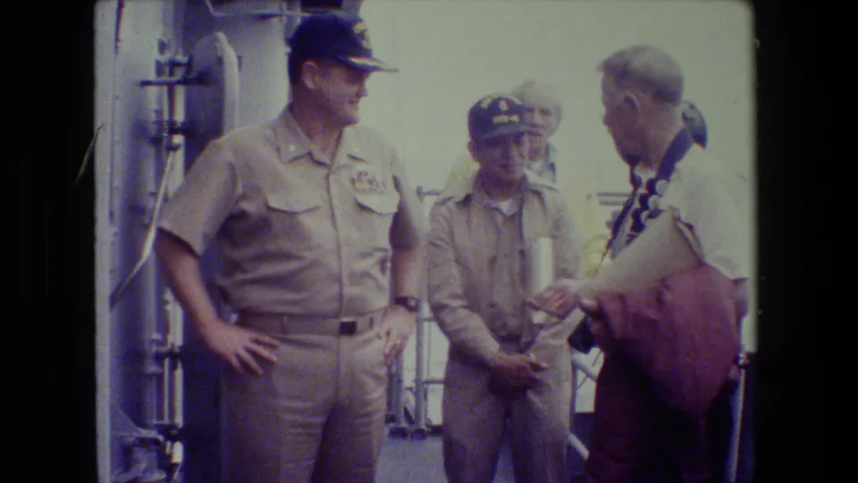 A khaki-uniformed naval officer stands with hands on hips beside another uniformed man and a civilian visitor holding camera gear on a ship deck.