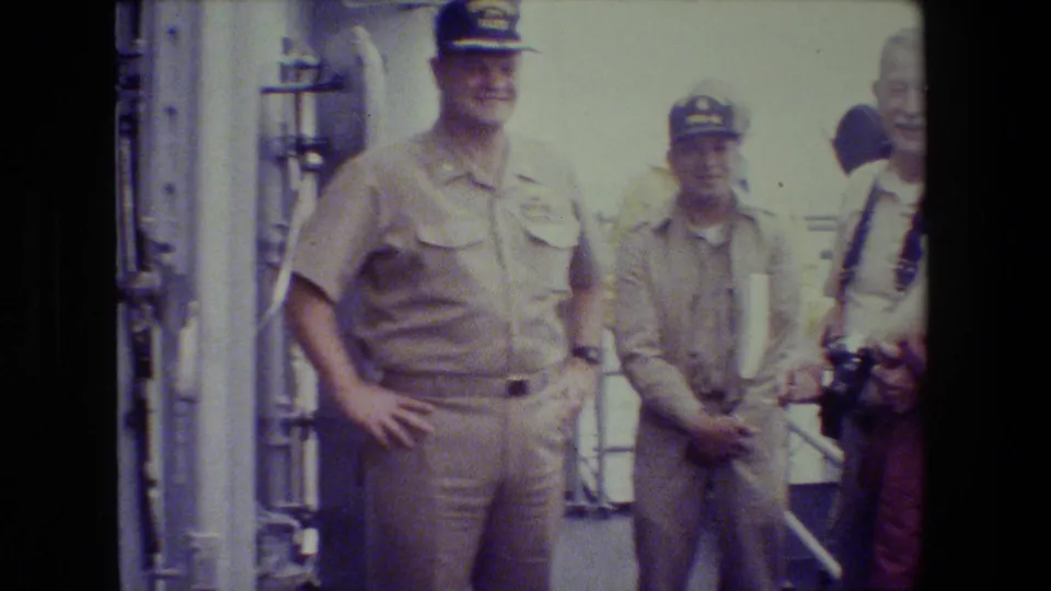 A khaki-uniformed officer and another uniformed man stand on the deck while a civilian visitor with camera gear is partly cropped at the right edge.