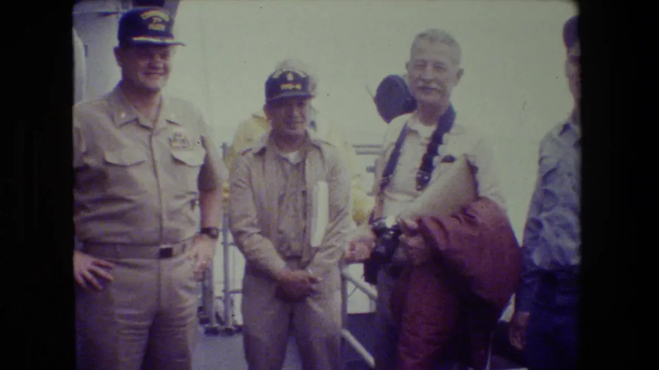 Uniformed men and a civilian visitor with cameras stand together on a bright ship deck, with another person partly visible at the edge of the frame.
