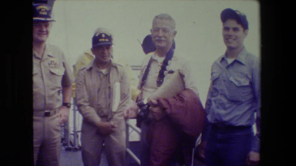 Early 1980s home-movie frame of naval personnel and a younger man posing on a boat deck, with one man holding a camera and railings behind them.
