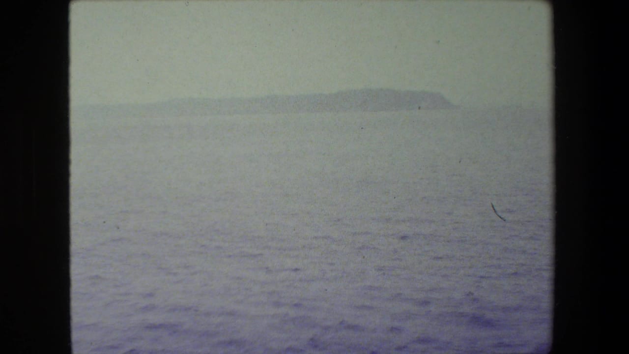 Hazy 8mm view across open sea toward a low distant island or headland, with no people visible in this frame.