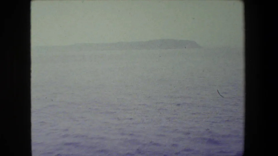 Hazy 8mm view across open sea toward a low distant island or headland, with no people visible in this frame.