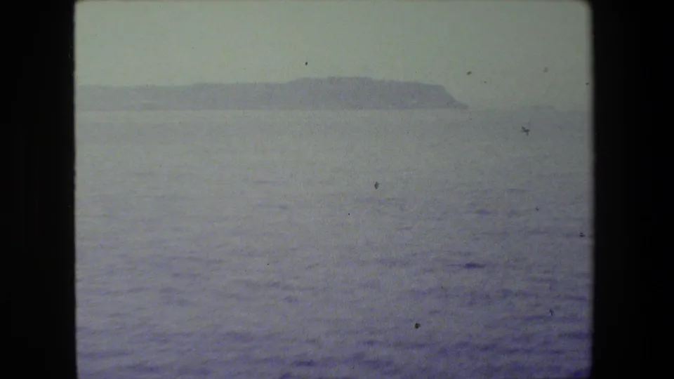 Soft, faded 8mm frame showing rippled ocean water and a low distant island or headland on the horizon.