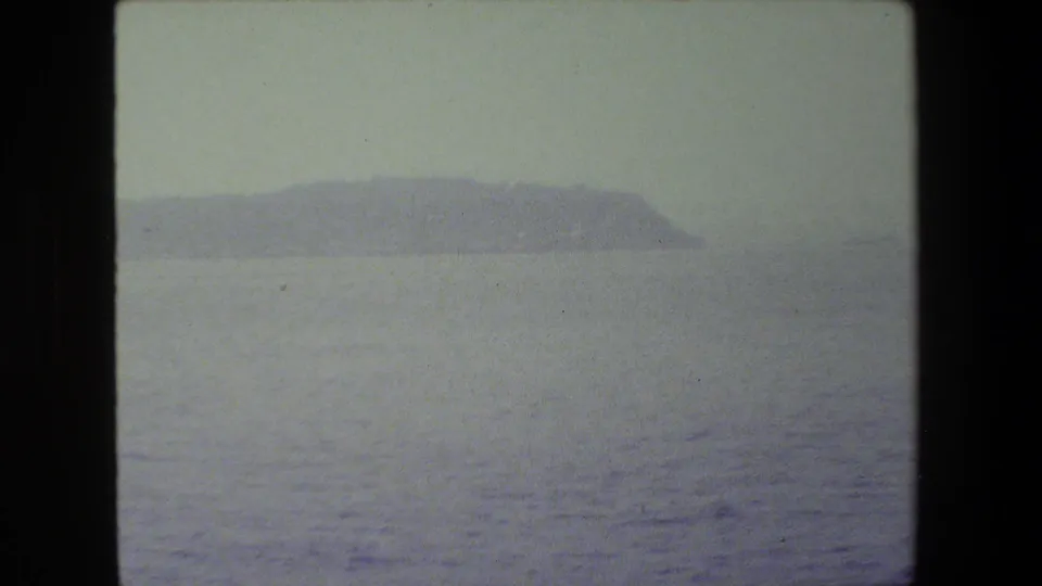Faded home-movie frame of open sea with a hazy, low island or headland stretching across the distant horizon.