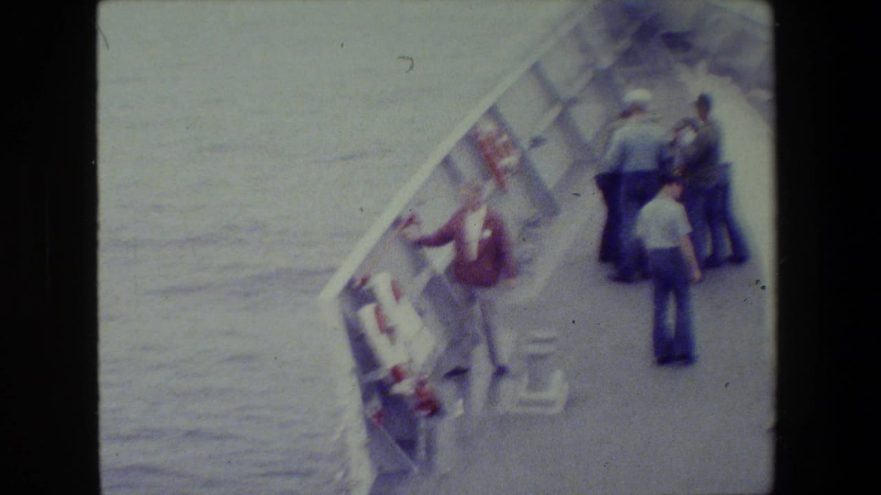 1981 archival 8mm footage shows several men on a naval vessel deck, with one man in a red jacket leaning over the side near a life ring and open water.