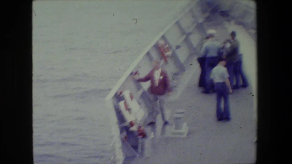 1981 archival 8mm footage shows several men on a naval vessel deck, with one man in a red jacket leaning over the side near a life ring and open water.