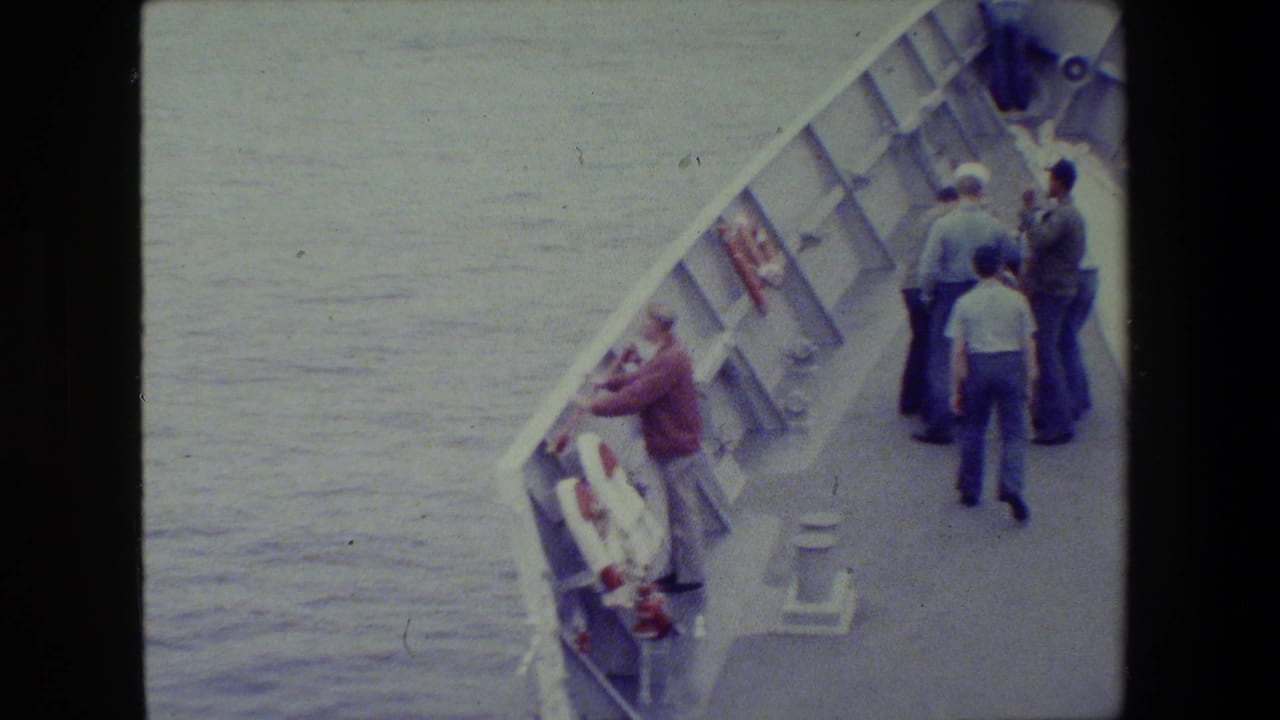 A man leans over a vessel railing while several men stand together farther along the deck.