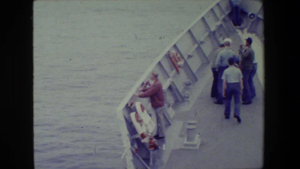 Several men are visible on the deck of a vessel in 1981 footage, with open water at left and a life ring mounted near a man leaning over the railing.