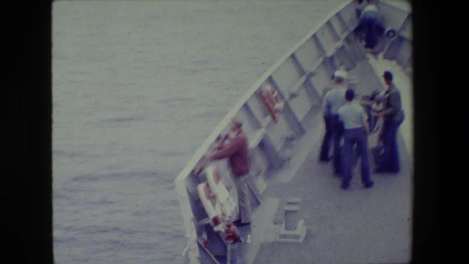 The frame shows a shipboard deck scene with men standing near the rail, a life ring mounted along the side, and water visible beyond the vessel.