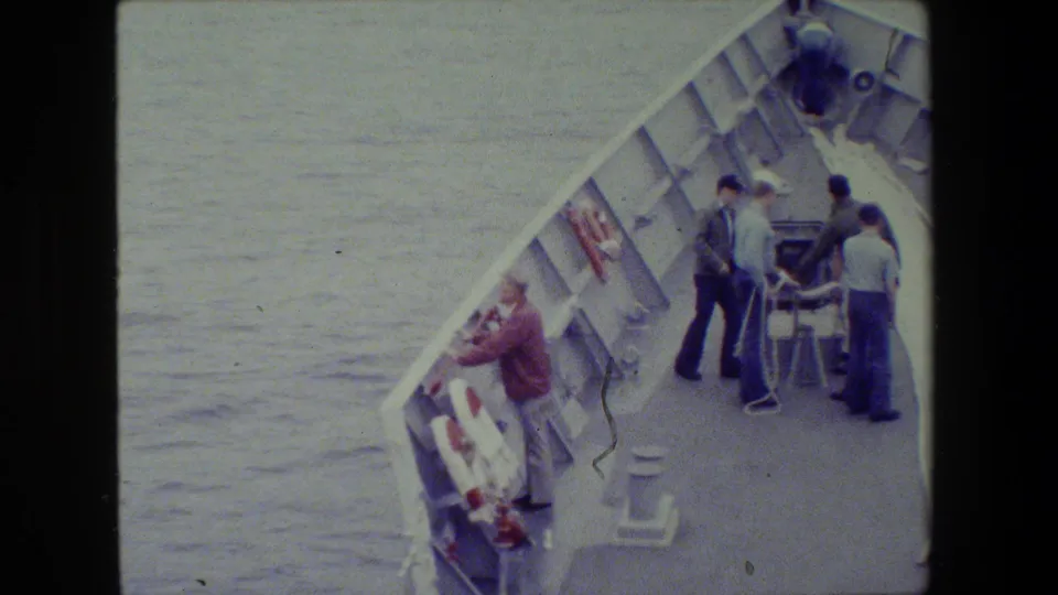 Near the end of the clip, several men gather around deck equipment on a naval vessel while a man in a red jacket remains near the side railing above the water.