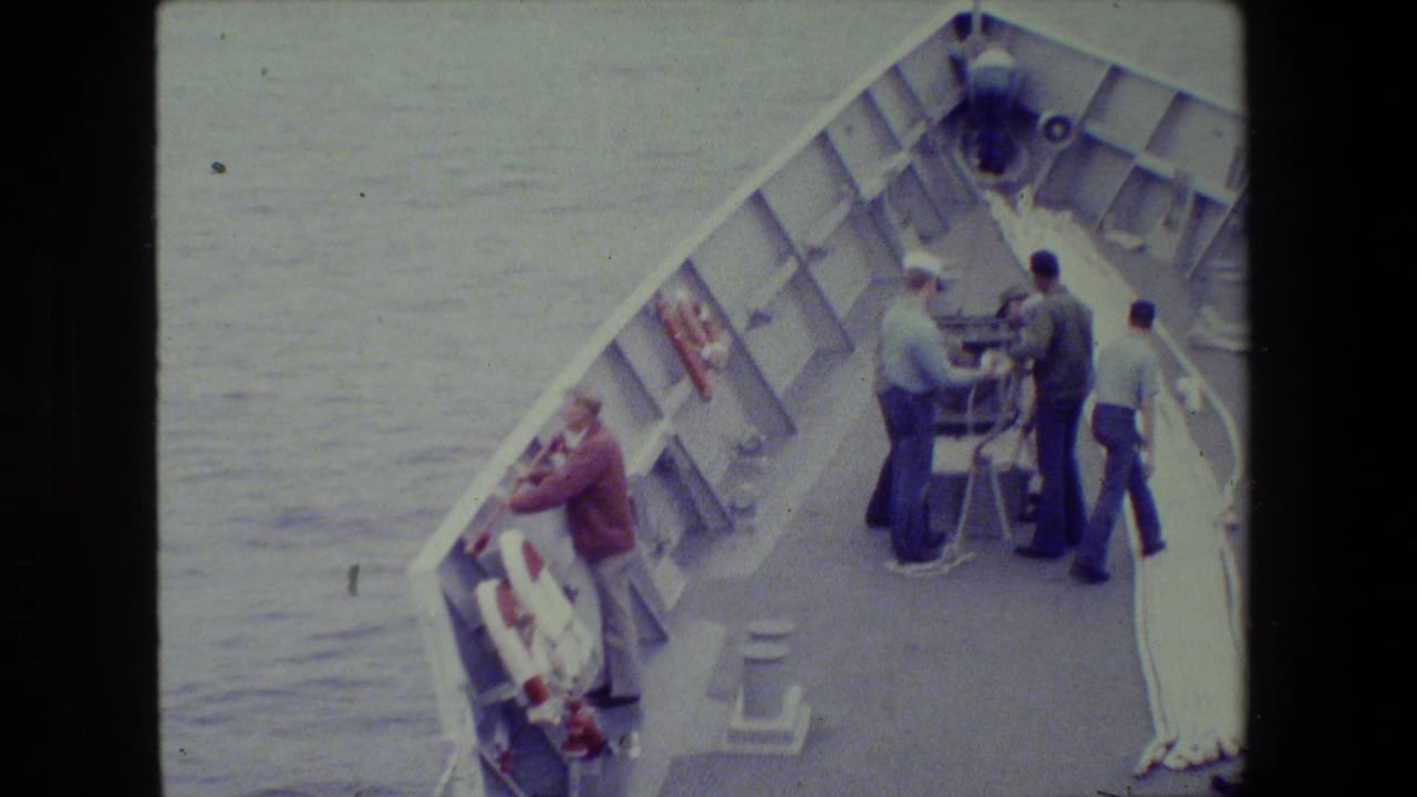High-angle 8mm frame of several adults on a ship's forward deck at sea, with railings, lines, deck fittings, and open water visible.