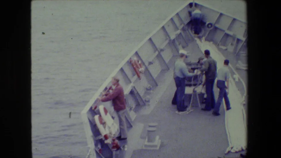 High-angle 8mm frame of several adults on a ship's forward deck at sea, with railings, lines, deck fittings, and open water visible.