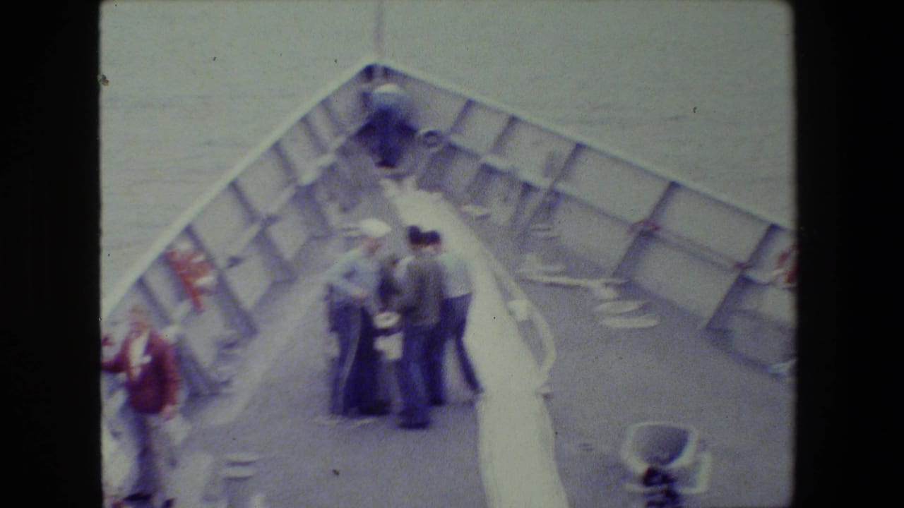 Soft 8mm frame showing adults on a vessel's bow area, some in uniform-like clothing, gathered around lines or equipment with the sea in the background.