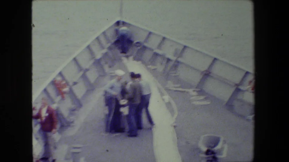 Soft 8mm frame showing adults on a vessel's bow area, some in uniform-like clothing, gathered around lines or equipment with the sea in the background.