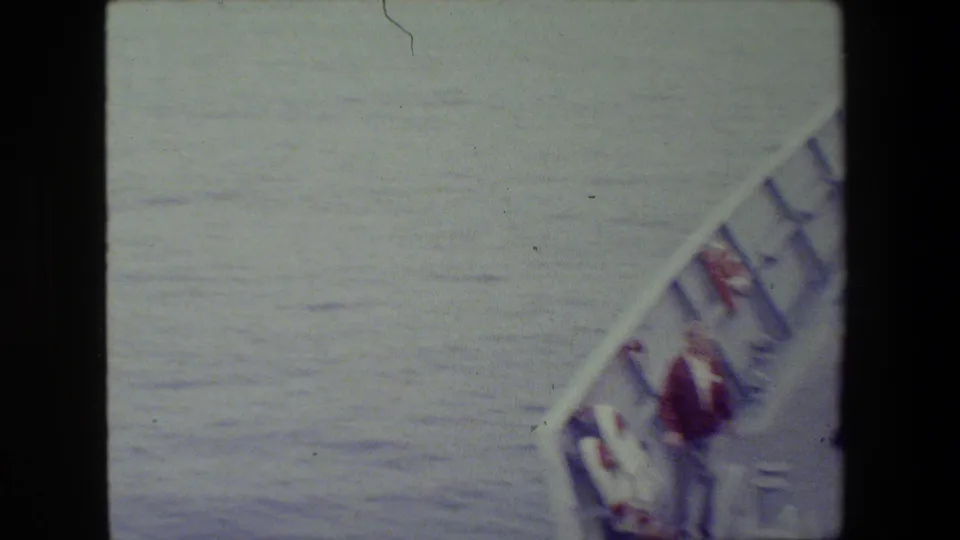 Blurred high-angle 8mm frame of the ship's forward rail and a small adult figure near the edge, with open water dominating the view.