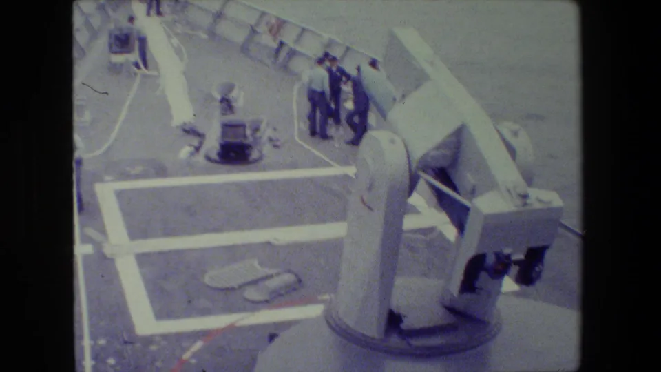 1981 8mm footage shows uniformed sailors on a naval ship deck near a large gun turret, railings, ropes, and painted deck markings.