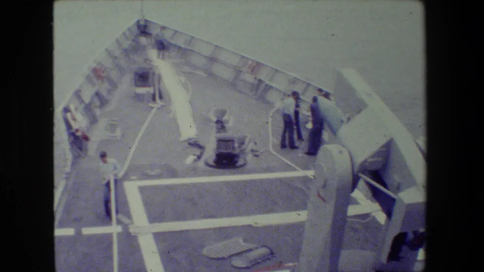 1981 8mm frame shows a naval vessel deck from above, with sailors near the railing and a large gun turret in the foreground.