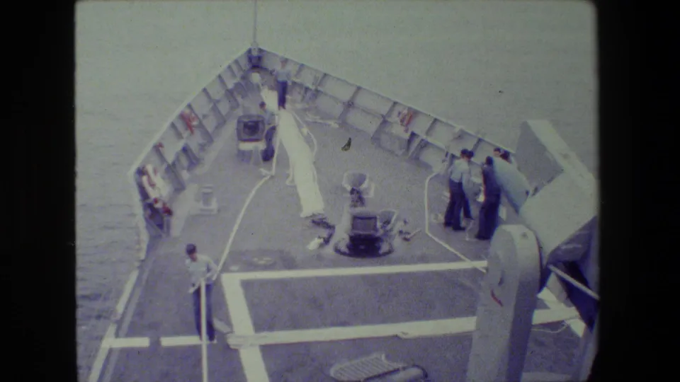From an elevated angle, sailors on a military ship's foredeck appear to work around a long white hose while open water surrounds the vessel.