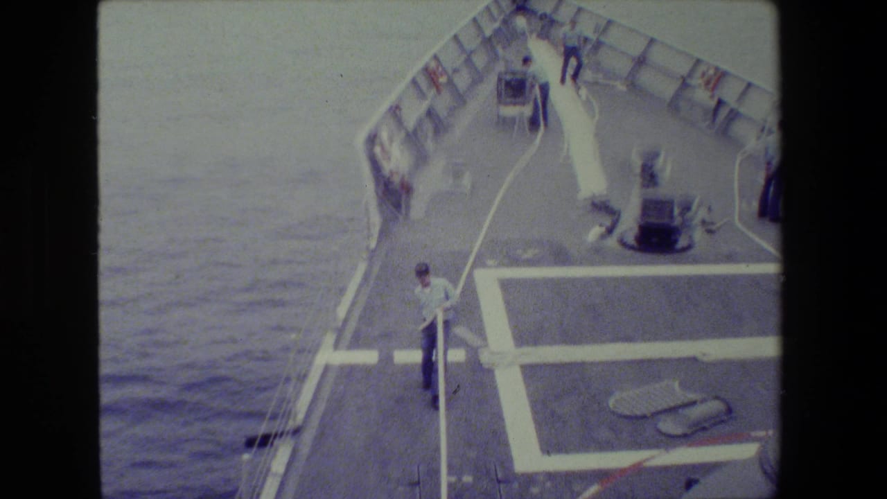 Ship bow with sailors, a white hose, deck markings, railings, and ocean on both sides.