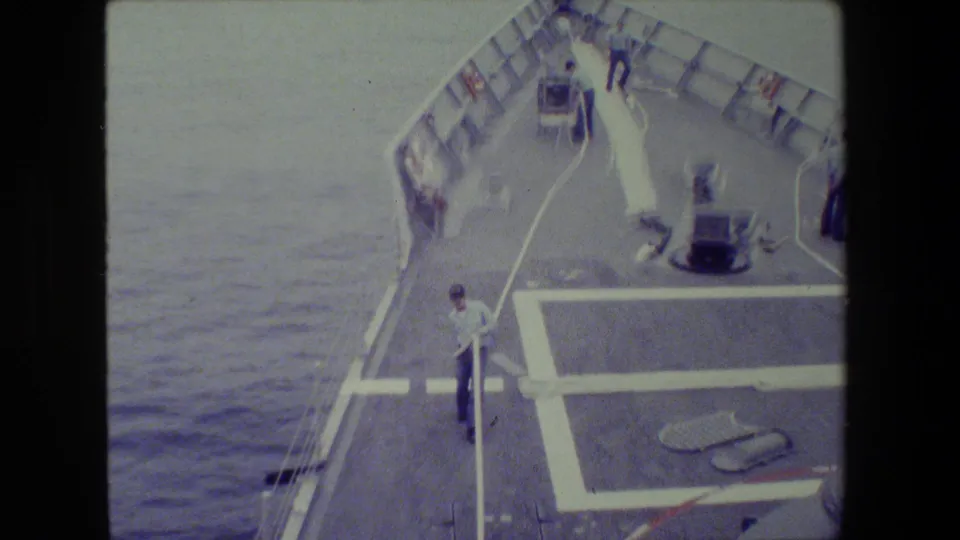 The ship's bow deck is seen from above with sailors near a long white hose, white deck markings, railings, and surrounding ocean.