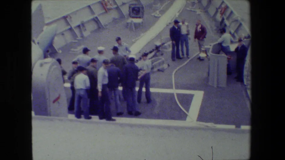 A larger group of uniformed sailors gathers on the deck near equipment while others stand by the long white hose on the ship at sea.
