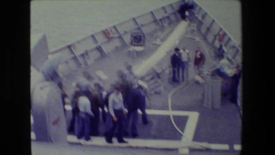 A slightly blurred handheld frame shows sailors grouped on the foredeck near a long white hose, deck railings, painted lines, and open water.