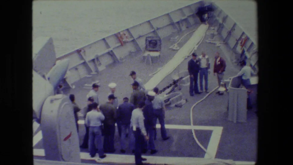 A high-angle 8mm frame shows sailors and other men on a grey naval vessel deck, clustered near a large launcher-like structure and white deck markings.