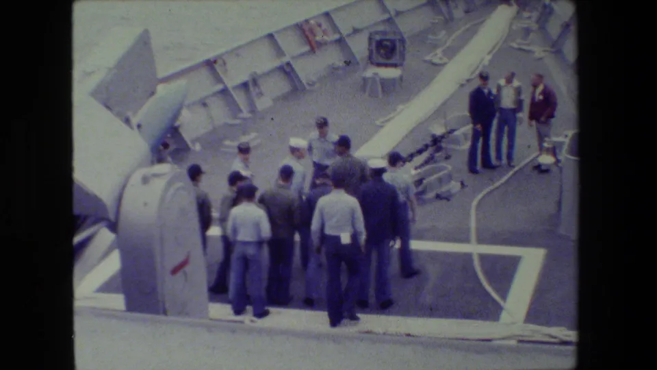 Sailors and men in work clothes stand in two loose groups on a naval deck, with a large equipment housing in the foreground and water visible beyond the rail.
