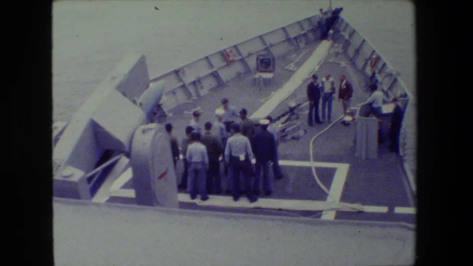 The frame shows a handheld 8mm view of sailors and other men gathered around shipboard hardware, with a large launcher-like object angled along the deck.