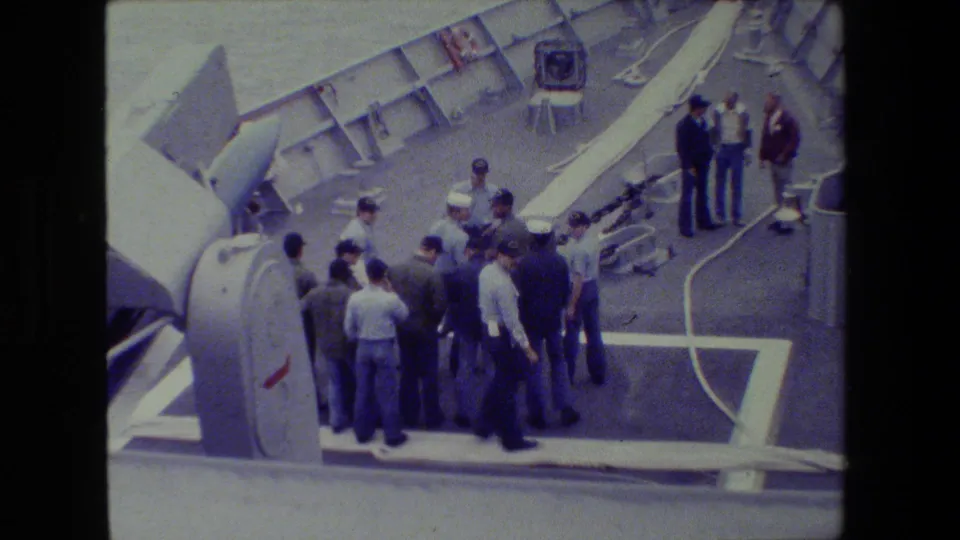 A group of sailors and men in work clothing remain gathered on the grey deck near large naval equipment, with white painted markings and the vessel edge visible.