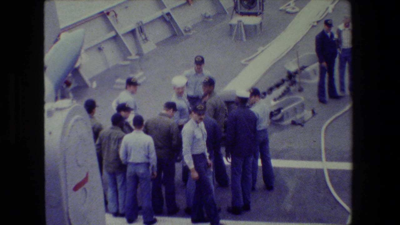 A group of uniformed naval personnel and civilian men stand on a ship deck, with chains, railings, and deck fittings visible in faded 8mm footage.