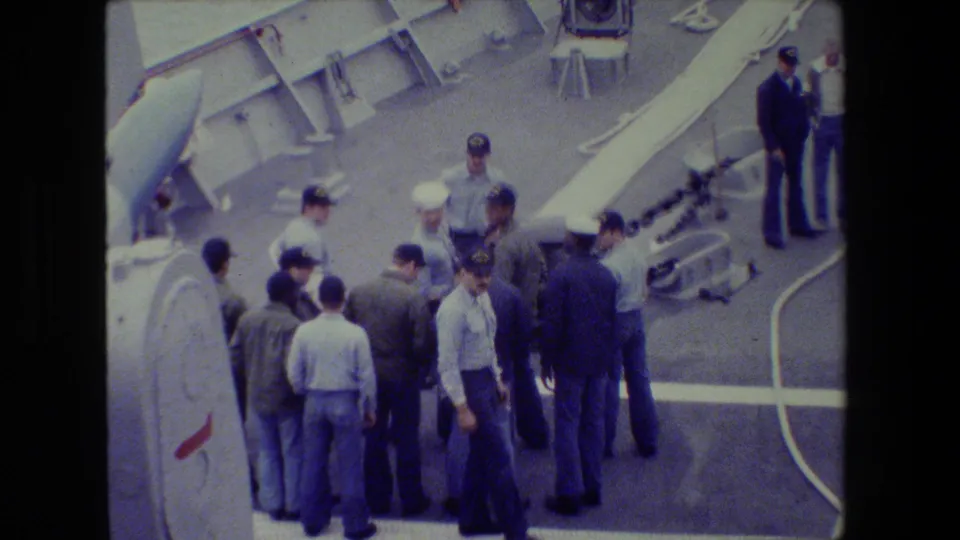 A group of uniformed naval personnel and civilian men stand on a ship deck, with chains, railings, and deck fittings visible in faded 8mm footage.