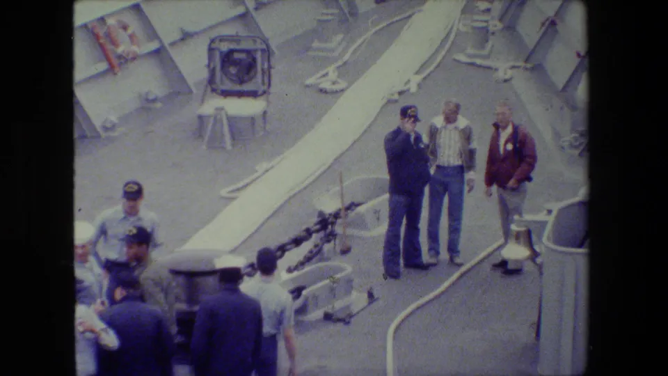 Several men, including uniformed personnel and civilians, stand on a naval ship deck and appear to look out over the water beyond the frame.