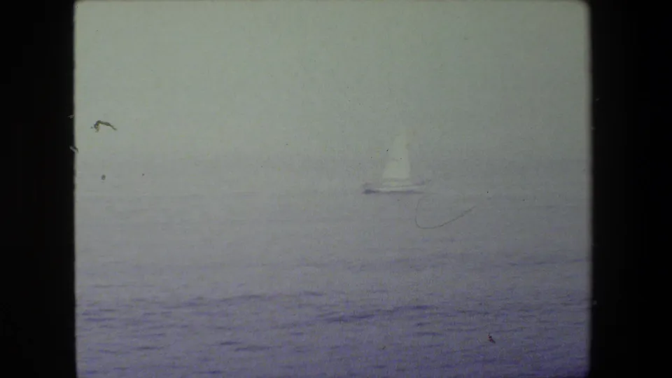 A distant sailboat moves across open water in a soft, hazy 8mm long shot, with the horizon and sea surface faintly visible.