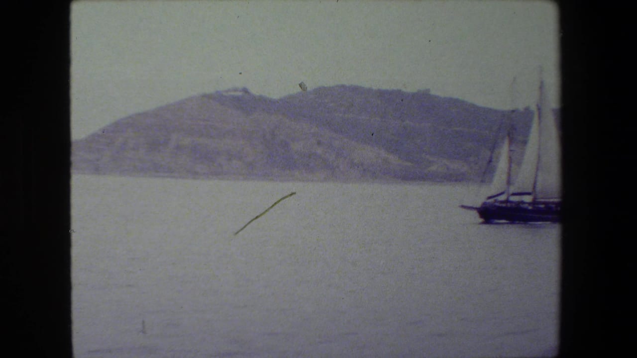 A dark-hulled sailboat with tall masts begins to enter from the right side of an 8mm frame, crossing calm water in front of a hilly shoreline.
