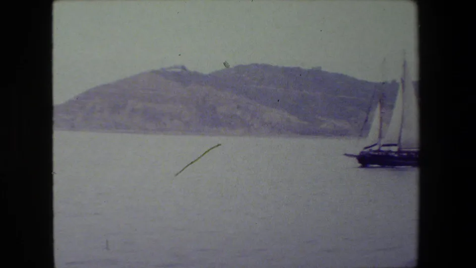 A dark-hulled sailboat with tall masts begins to enter from the right side of an 8mm frame, crossing calm water in front of a hilly shoreline.
