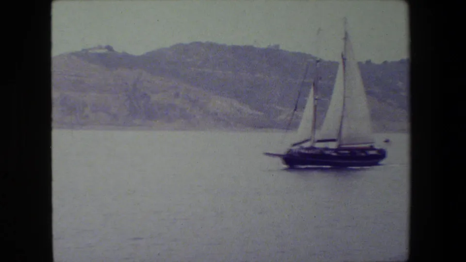 The sailboat is more centered in the frame, with white sails raised above a dark hull as it moves across calm water against a soft hilly shoreline.