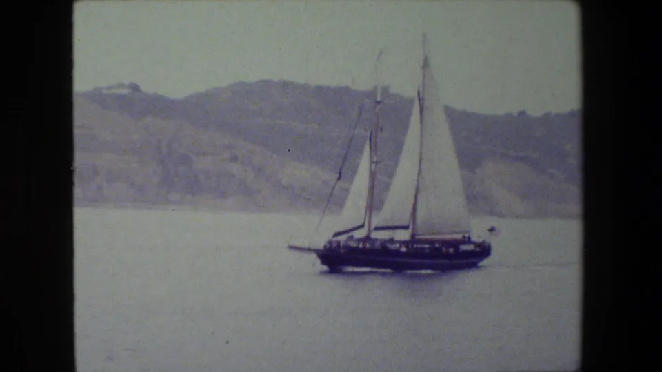 A dark-hulled sailboat with multiple masts and white sails sits prominently on calm water, framed by distant hills and the soft texture of 8mm home-movie film.