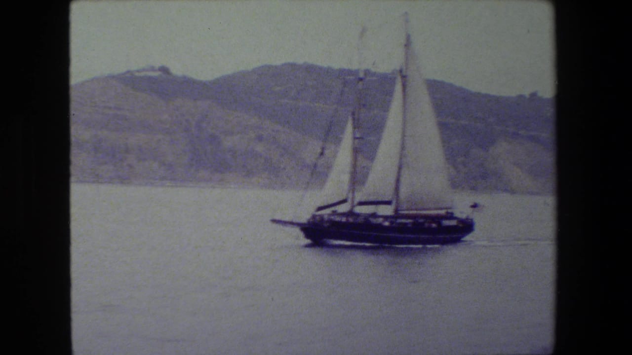 A dark-hulled sailboat with raised white sails moves across calm water, with a hazy hillside and pale sky in the background.