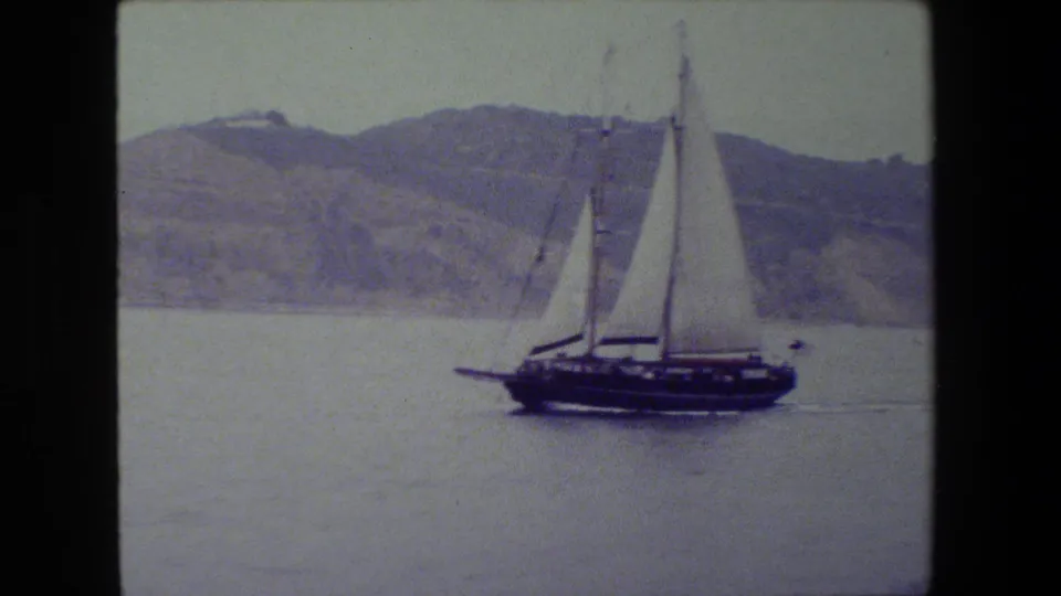 A dark-hulled sailboat with raised white sails moves across calm water, with a hazy hillside and pale sky in the background.